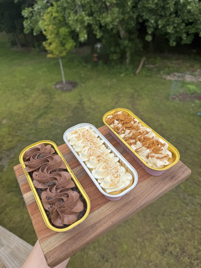 Three mini cakes in tins — chocolate, coconut, and caramel — on a wooden board outdoors